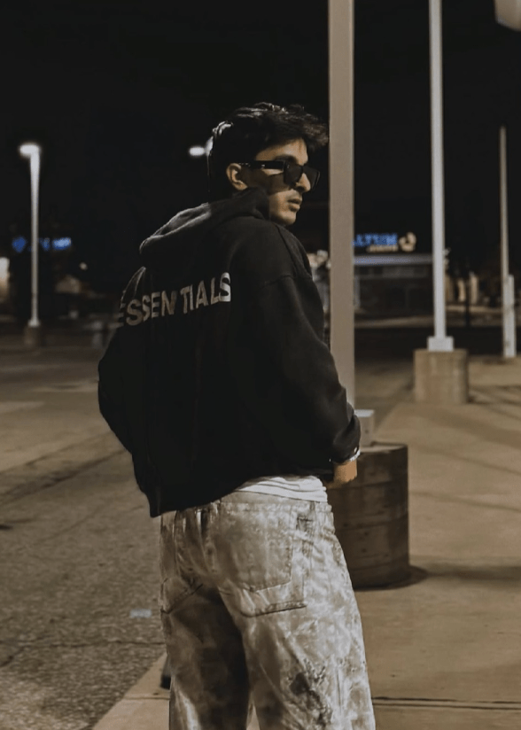 An Indian boy wearing a black hoddies with glasses, you can see his back towards the camera and essentials written on the back of his hoodie